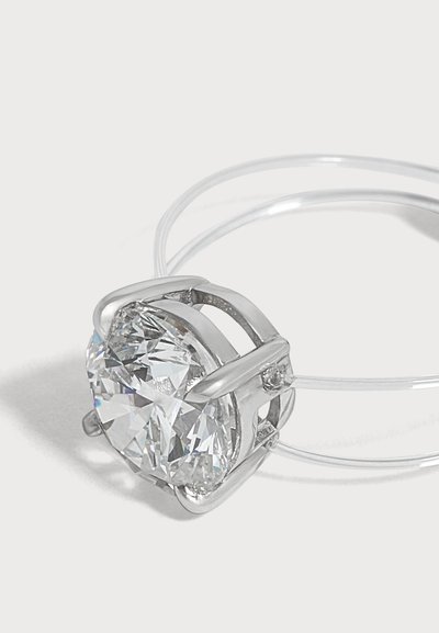 Anillo de plata con una gran piedra preciosa facetada, clara y redonda, sostenida por cuatro garras sobre una banda fina y transparente, sobre un fondo blanco.