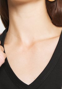 Černý top s výstřihem do V a hladkou texturou, zdobený kontrastním prošitím. Na obrázku je částečně vidět světle růžový rukáv svetru.