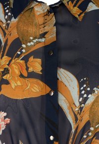 Blusa floreale con sfondo scuro con fiori arancioni e gialli, colletto ordinato e chiusure con bottoni. Tessuto leggero con una texture liscia.