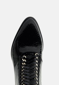Botine negre din piele cu vârf ascuțit, prezentând un finisaj texturat, un design complicat de șireturi și ochiuri metalice.