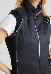 Femme portant un gilet matelassé sans manches noir et gris foncé avec une fermeture éclair, par-dessus une chemise blanche à manches courtes, se tenant avec les mains sur les hanches.