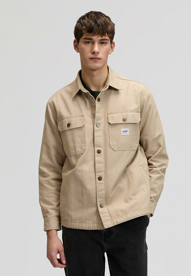 Jeune homme portant une veste beige à boutons avec des poches avant et un patch logo Lee visible, assortie à un pantalon noir, debout devant un fond uni.