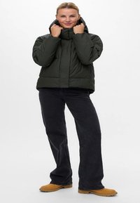 Mørk grønn puffjakke med høy krage og delvis glidelås, båret over svarte vide jeans. Brune sko med teksturerte såler.