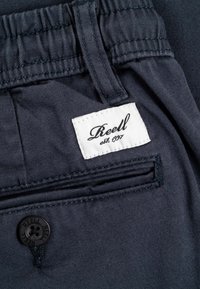 Los pantalones de algodón azul marino presentan una etiqueta blanca con la inscripción "Reell est. 1997." Se observa un botón negro y un detalle de bolsillo trasero cosido.