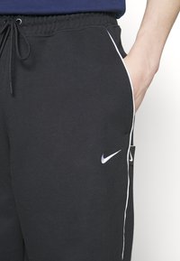 Persona con pantalones deportivos Nike negros con ribete blanco, mano parcialmente en el bolsillo derecho y cintura elástica con cordón.