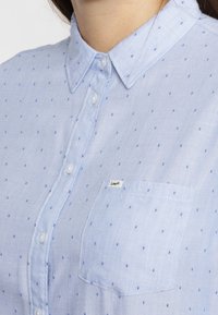 Hellblaues Button-up-Hemd aus strukturiertem Stoff, mit kleinen dunkleren blauen Punkten, einer Brusttasche und einem Kragen mit Kunststoffknöpfen.
