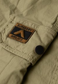 Olijfgroen stof met een gladde textuur, voorzien van een bruine en zwarte geweven patch met de tekst "Superdry" en een schuine vorm. Zwarte knoopaccent.