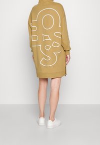 Huvtröjklänning i senapsgul med en stor vit grafik på baksidan, med siddragkedjor och långa ärmar, i kombination med vita sneakers.