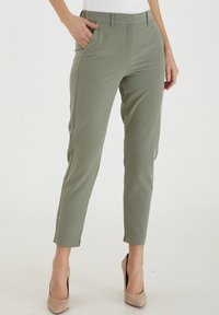 Kvinna i slim-fit, korta olivgröna byxor med sidofickor och beige högklackade skor, stående på ett vitt golv.