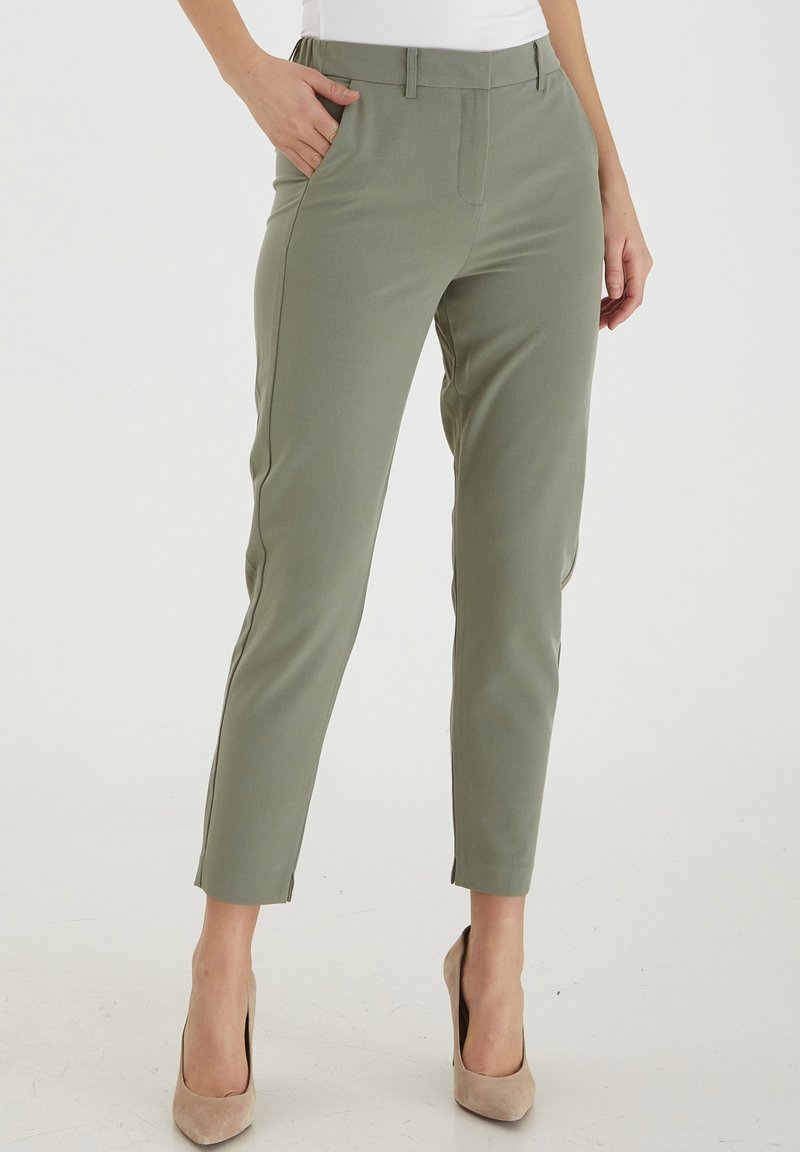 Kvinna i slim-fit, korta olivgröna byxor med sidofickor och beige högklackade skor, stående på ett vitt golv.