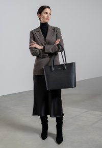 Sac fourre-tout noir texturé avec deux poignées, associé à un blazer marron sur un col roulé noir et une jupe en satin noir.