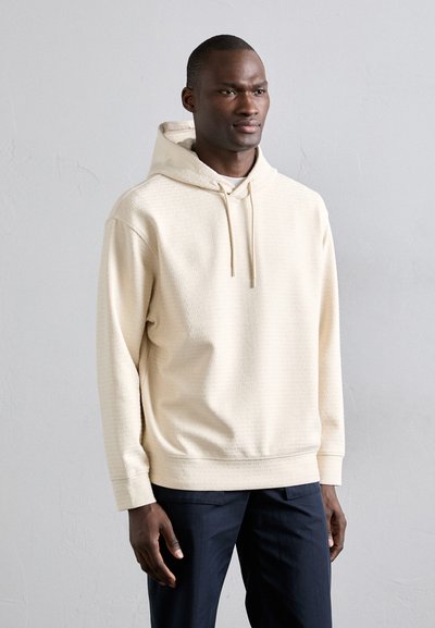 Sweatshirt à capuche couleur crème en tissu texturé avec une poche kangourou. Présente des cordons de serrage et des poignets côtelés. Porté avec un pantalon sombre.
