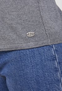 Grå sweater med en struktureret overflade, der har et lille metaltag med "edc since 1986" ved kanten. Kombineret med blå denimstof.