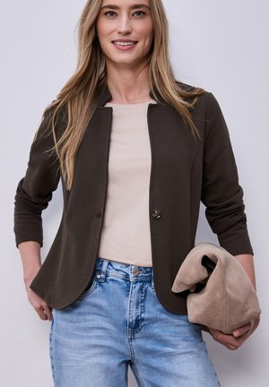 Frau lächelt, trägt eine dunkelbraune Jacke über einem beigen Oberteil mit hellblauen Jeans und hält eine gefaltete beige Wildledertasche in der rechten Hand.
