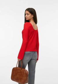 Vrouw met lang donker haar, gekleed in een rode, gestructureerde, lange mouwen top en grijze spijkerbroek, met een bruine leren handtas in de hand, kijkend over haar schouder.