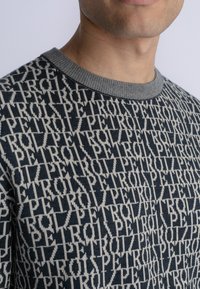 Pulover tricotat de culoare bleumarin, cu un model de text repetat în crem. Gulerul rib accesorizat adaugă textură și contrast designului.