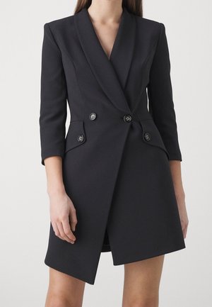 Kobieta w czarnej, dwurzędowej sukience marynarkowej z rękawami do połowy przedramienia i asymetrycznym dołem na jednolitym tle.