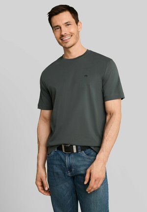 Homme souriant portant un T-shirt gris foncé à manches courtes et un jean bleu avec une ceinture noire, debout devant un fond uni gris clair.