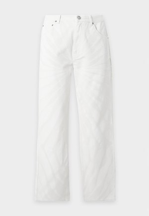 Homme portant un haut sans manches blanc transparent à motif, un pantalon blanc ample et des chaussures habillées noires et blanches, debout devant un fond uni.