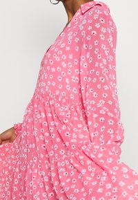 Abito floreale rosa con colletto, maniche lunghe e gonna a balze. Il tessuto presenta piccoli fiori bianchi e rossi, texture leggera e fluida.