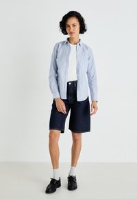 Ανοιχτό μπλε ριγέ πουκάμισο με κουμπιά, λευκό μπλουζάκι από κάτω, σκούρα τζιν σορτς, μαύρα παπούτσια, γκρι κάλτσες, στέκεται σε απλό φόντο.