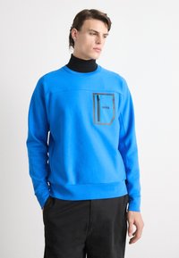 Uomo che indossa un felpa blu brillante con tasca centrale con zip etichettata "BOSS" sopra un dolcevita nero e pantaloni neri, con le mani in tasca.