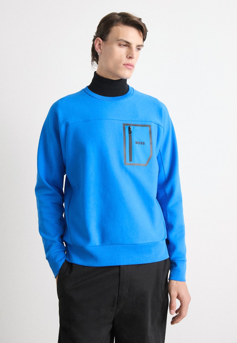 Uomo che indossa un felpa blu brillante con tasca centrale con zip etichettata "BOSS" sopra un dolcevita nero e pantaloni neri, con le mani in tasca.