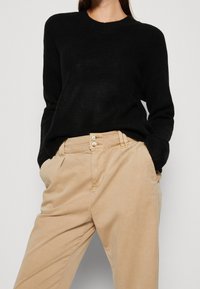 Personne portant un pull noir à manches longues rentré dans un pantalon beige taille haute avec deux boutons, les mains dans les poches.