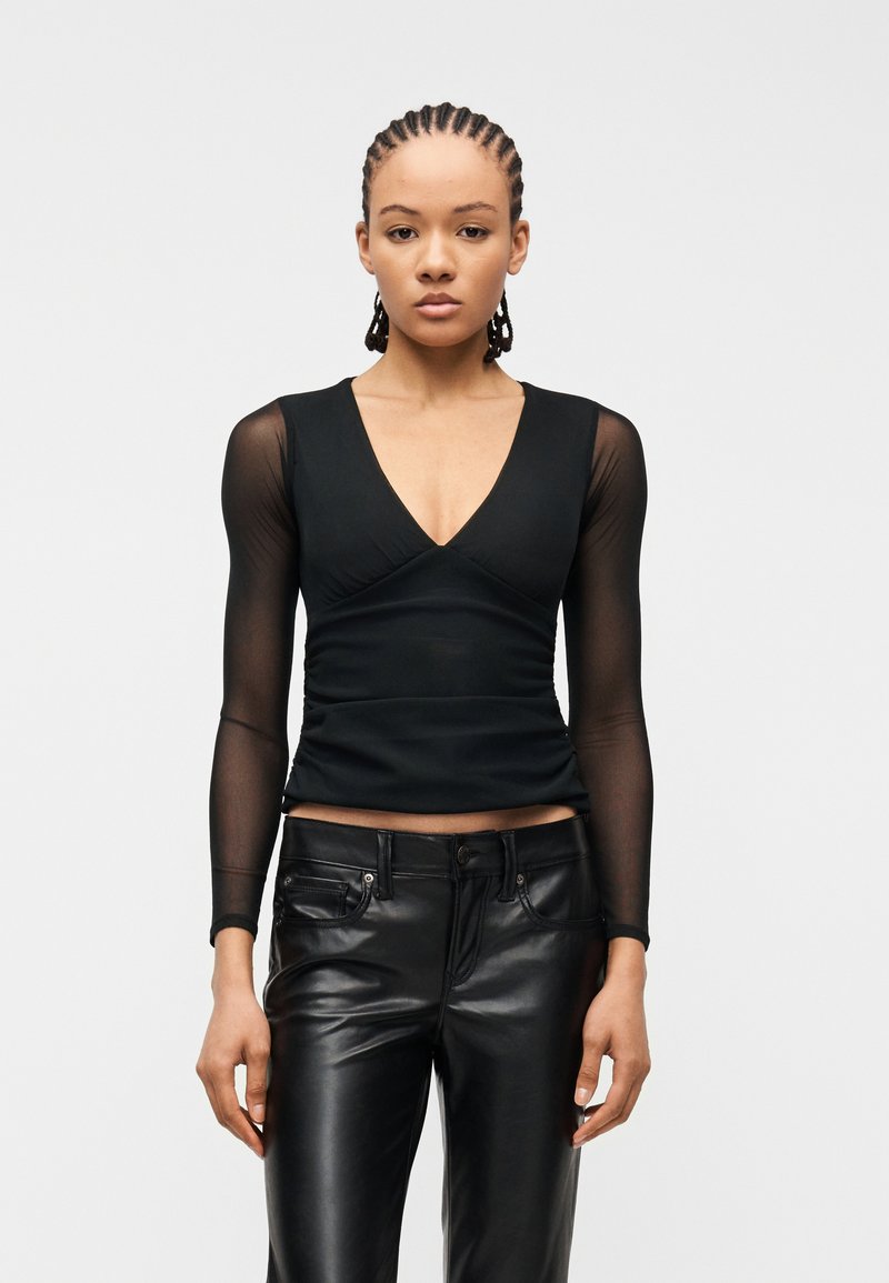 Νεαρή γυναίκα με πλεξούδες στα μαλλιά, που φοράει μαύρο τοπ με V λαιμόκοψη και διάφανα μακριά μανίκια, καθώς και μαύρο δερμάτινο παντελόνι, κοιτάζοντας μπροστά πάνω σε λευκό φόντο.