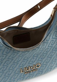 Borsa a tracolla in denim blu con chiusura a zip, ferrature dorate, logo impresso e tracolla in pelle marrone liscia. Motivo superficiale strutturato visibile.