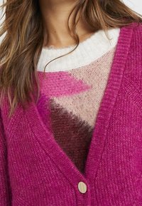 Femme portant un pull douillet multicolore sous un cardigan tricoté rose vif boutonné, montrant la poitrine et le cou.