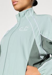 Αντιανεμικό ελαφρύ σε μέντα και σκούρο πράσινο, με ψηλό γιακά, κλείσιμο με φερμουάρ και λευκές διακοσμητικές γραμμές. Διακρίνεται το λογότυπο Champion.