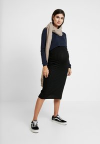 Conjunto de maternidade com uma blusa de manga longa azul-marinho, saia midi preta com textura e ténis pretos. Um lenço bege envolve o pescoço.