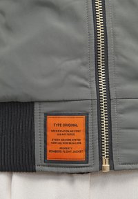 Giacca da volo bomber caratterizzata da un tessuto grigio, orlo nero a coste e un'etichetta arancione prominente con specifiche e dettagli del marchio.