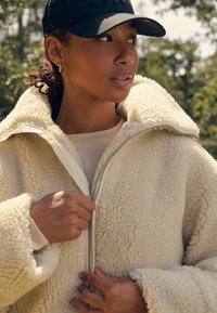 Kremīgu shearling jaku ar augstu apkakli, ar rāvējslēdzēju un teksturētu audumu. Modelis valkā gaišu augšu un melnu cepuri.