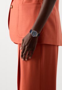 Marinblå klocka med åttakantig urtavla, silverdetaljer och en texturerad rem, bärs på en hand mot en orange kavaj.