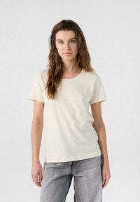 Camiseta de manga corta de color crema, hecha de tela suave, con un escote redondeado y un sutil acento de logo cerca del dobladillo.