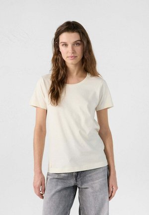 Camiseta de manga corta de color crema, hecha de tela suave, con un escote redondeado y un sutil acento de logo cerca del dobladillo.