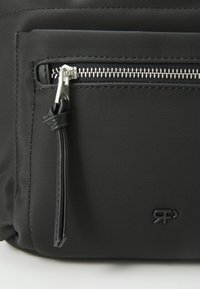 Gros plan sur une poche d'un sac en tissu noir avec une fermeture éclair argentée et un tirette en cuir, arborant un petit logo noir embossé.