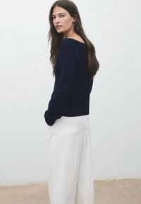 Femme aux cheveux bruns longs portant un haut à manches longues bleu marine et un pantalon large blanc, se tenant devant un mur blanc uni.