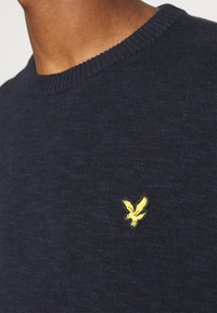 Närbild av en marinblå stickad tröja med en gul broderad fågel-logotyp på bröstet nära halsringningen.