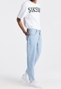 Camiseta blanca de manga corta con la palabra "SIKSILK" impresa en negro, combinada con jeans de mezclilla azul claro y zapatillas blancas. Corte recto.