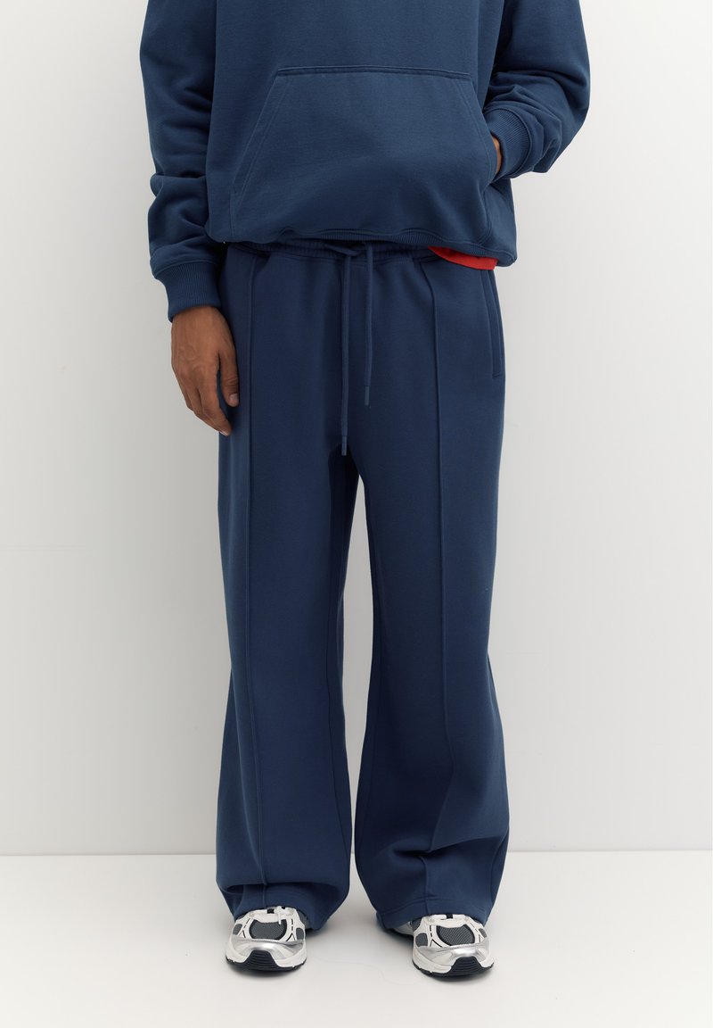 Personne portant un pantalon de survêtement large bleu marine, un sweat à capuche bleu marine assorti et des baskets blanches, debout devant un fond clair uni.
