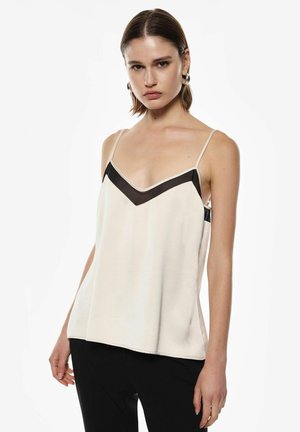 Giovane donna che indossa una canotta beige senza maniche con dettaglio a zigzag nero e pantaloni neri, posando davanti a uno sfondo bianco uniforme.