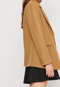 Hnědý strukturovaný blejzr s klopovým límcem, zapínáním na jeden knoflík a dlouhými rukávy; hladká textura; stylizovaný na černé sukni.