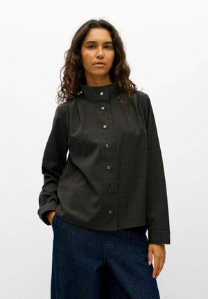 Un mannequin, une main dans la poche, porte une veste sombre à carreaux, boutonnée et à col montant, ainsi qu’un jean bleu foncé sur fond blanc.