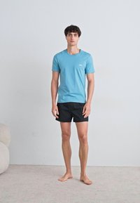 Lyseblå bomull T-skjorte med korte ermer og en liten logo på brystet, parret med svarte shorts. Barføtt, stående mot en nøytral bakgrunn.