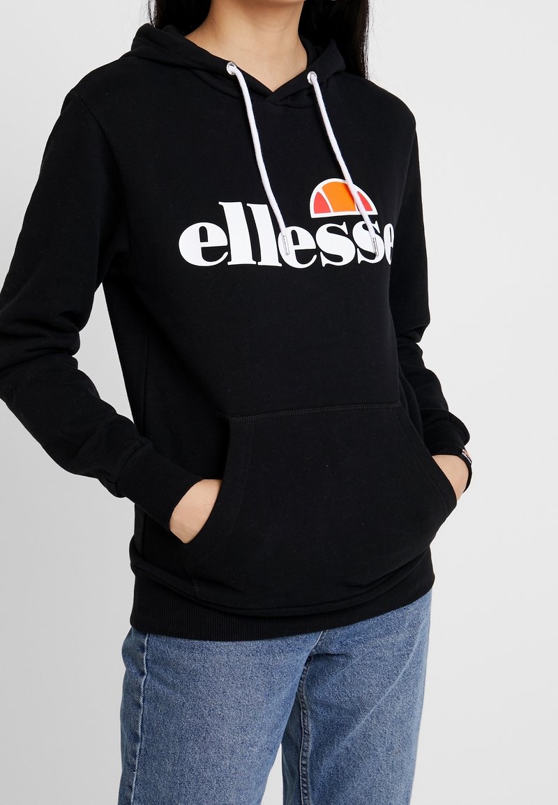 Černá mikina s kapucí a bílými šňůrkami, na které se nachází velké bílé logo "ellesse" a barevná grafika západu slunce. Přední klokaní kapsa a žebrovaný lem.