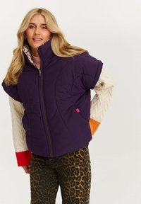 Fioletowa pikowana kurtka z krótkimi rękawami, zapinana na zamek, z różowymi detalami w kieszeni, w zestawie z teksturowanymi beżowo-pomarańczowymi dzianinowymi rękawami oraz spodniami w leopardzi wzór.