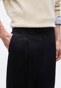 Personne portant un pull beige et un pantalon noir, main dans la poche, montrant les détails du devant plissé et de la ceinture.